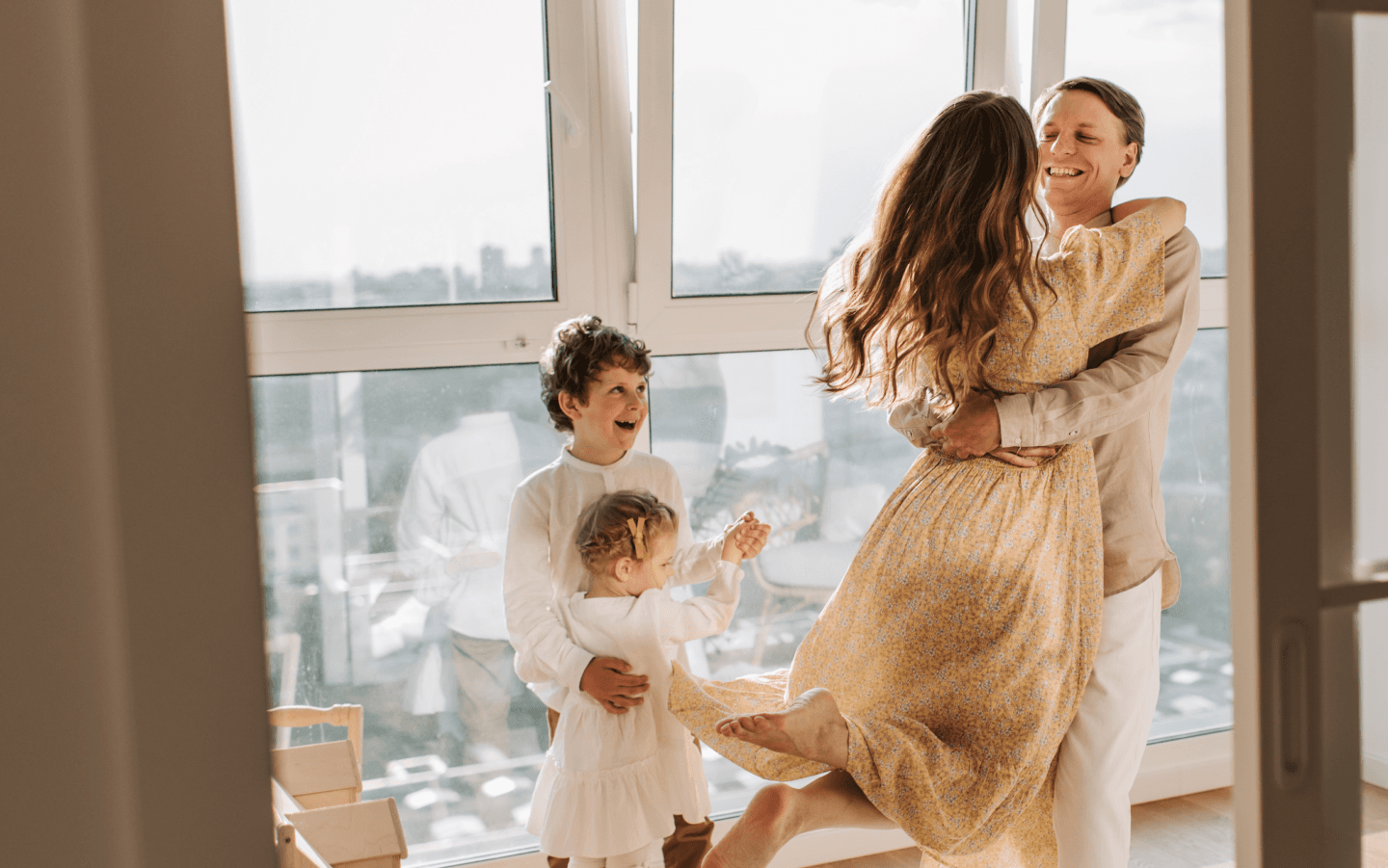 Familia feliz em casa com a luz do sol entrando pela janela e mãe e pai se abraçando com o filho e a filha sorrindo e brincando de dançar juntos.